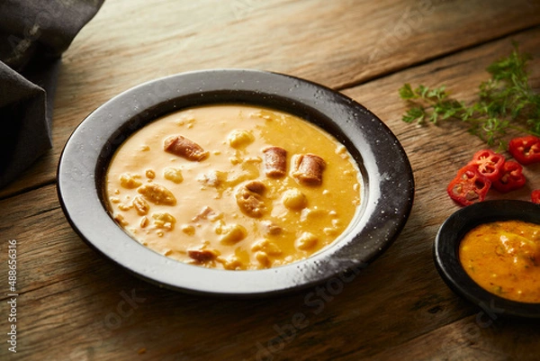 Fototapeta Mote pata is a traditional ecuadorian soup made from mote, pork, sausage and sambo seeds. It’s served on a white plate with a rustic, wooden and homey background. 