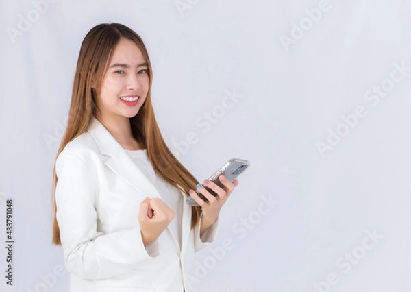 Obraz Beautiful professional Asian business woman in a white suit smiles happily successful while looking at the camera and holds smartphone.