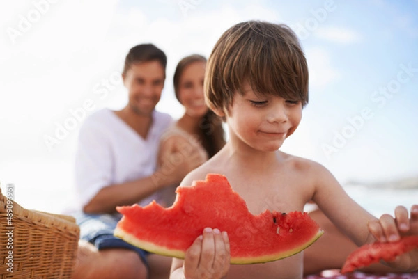 Obraz Juicy and sweet. A happy little boy biting into a watermelon with his parents looking on.