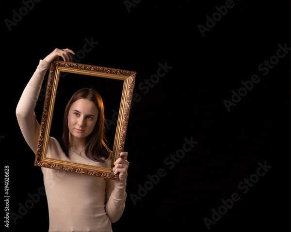 Fototapeta Get out of the comfort zone, beyond the psychological framework concept. A young woman holds a picture frame in her hands on a dark background.