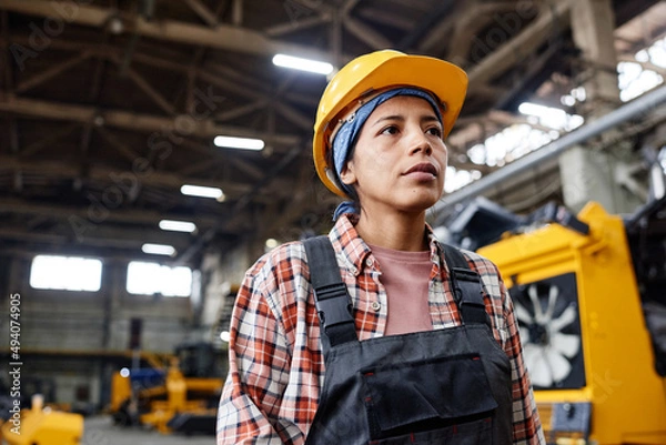 Fototapeta Young serious female worker of industrial plant wearing coveralls and hardhat standing in large distribution warehouse