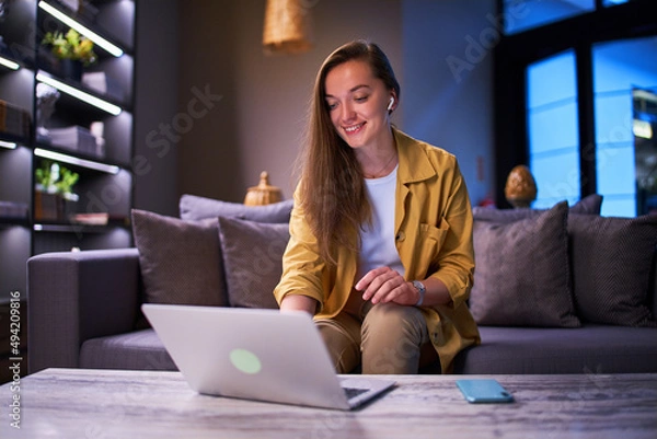 Fototapeta Happy cute joyful modern smiling satisfied smart young casual businesswoman wearing wireless headphones using laptop for working online at cafe