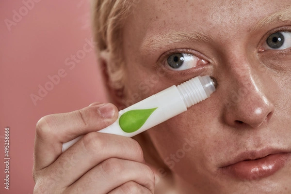 Fototapeta Guy applying depuffing eye serum on his face skin