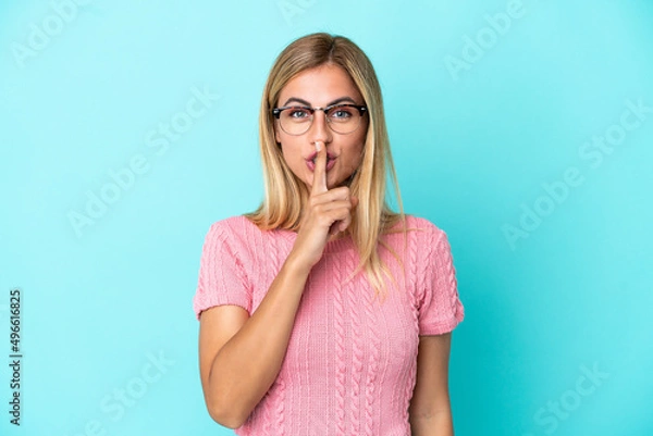 Fototapeta Blonde Uruguayan girl isolated on blue background showing a sign of silence gesture putting finger in mouth