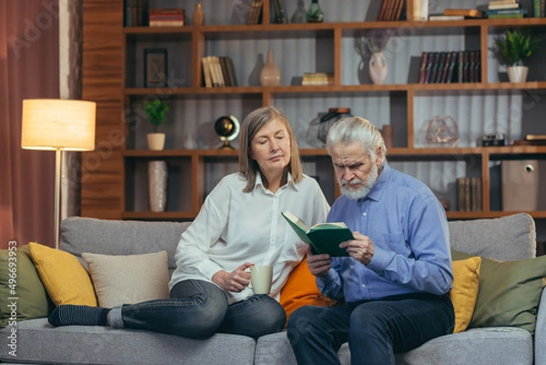 Fototapeta senior couple spending leisure time reading a book together sitting on cozy sofa at home. Elderly mature family Gray hair man and woman together carefree retirement lifestyle. Cozy in house