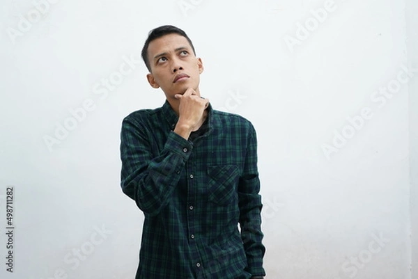 Fototapeta Portrait of a thinking young Asian man wearing casual clothes looking up isolated over white background