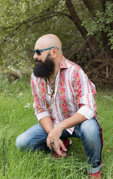 Fototapeta image of a bald man with beard using sunglasses posing on a green area