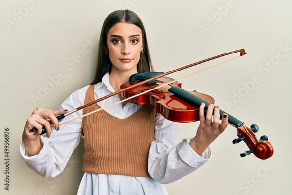 Fototapeta Beautiful brunette young woman playing violin relaxed with serious expression on face. simple and natural looking at the camera.