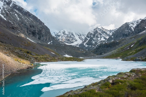 Fototapeta Atmospheric highland landscape with frozen alpine lake and high snowy mountains. Awesome scenery with icy mountain lake on background of large snow mountains in low clouds. Scenic view to ice lake.