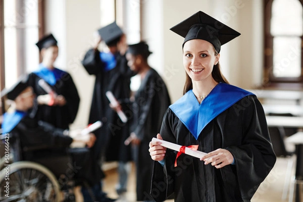 Fototapeta Waist up portrait of young woman wearing graduation gown and smiling at camera in university ceremony