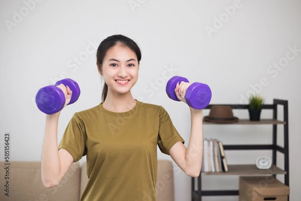 Fototapeta Attractive asian young fitness woman lifting dumbbell weights workout at home in living room. Fresh feeling female training and exercise wearing sport wear fit body.