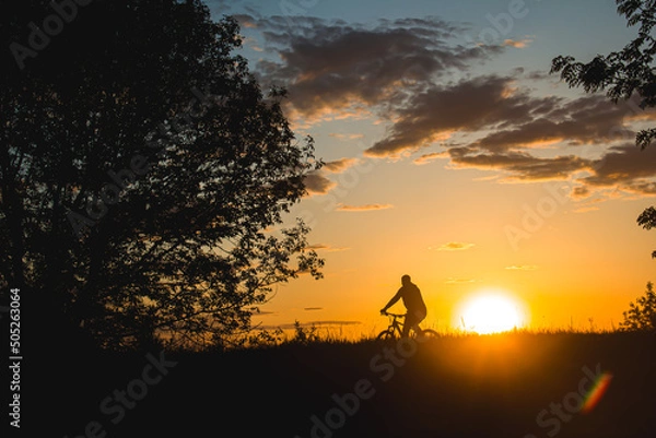 Fototapeta A cyclist rides through a field against the backdrop of a beautiful contour sunset light. Beautiful sunset. Photo of nature. Velotheme.