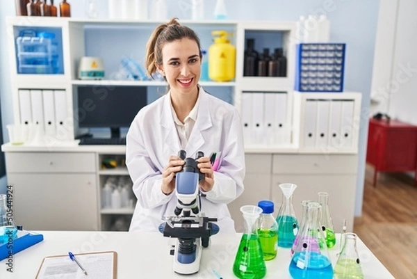 Fototapeta Young woman wearing scientist uniform using microscope at laboratory