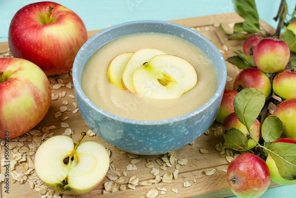 Fototapeta Baby's first complementary food, baby nutrition. Oatmeal porridge for the baby from ground cereals in a blue bowl, red ripe apples on a wooden board close-up.