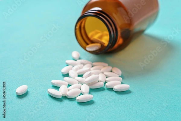 Fototapeta White oval medical pills pouring out of a medicine bottle on a blue background. Macro photo with copy space.