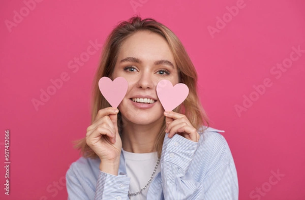 Fototapeta Medium close-up portrait of beautiful young woman with blond hair standing against pink wall background holding paper hearts smiling at camera