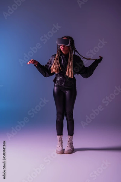 Fototapeta Full length of trendy african american woman using vr headset on blue and purple background.