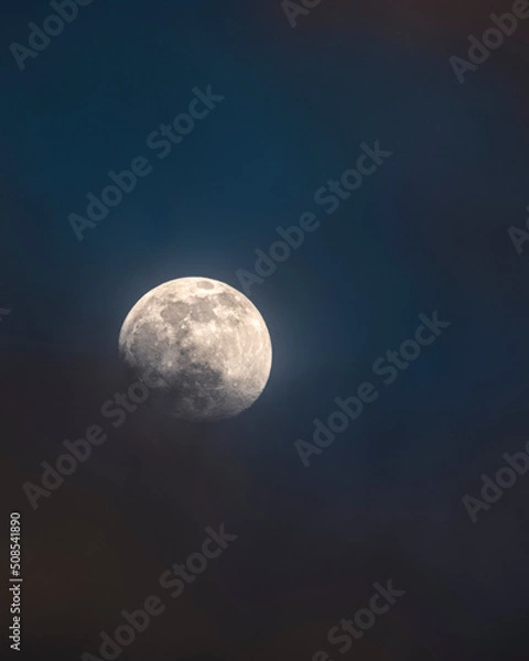 Fototapeta A super full Moon partially covered by clouds against a dark blue sky in the summer of 2022