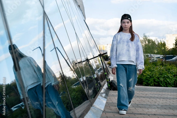 Fototapeta A teenage girl in jeans and a sweatshirt walks along a city street. The sky is reflected in the shop window. Blue background. Summer holidays in the city. Schoolchildren on vacation