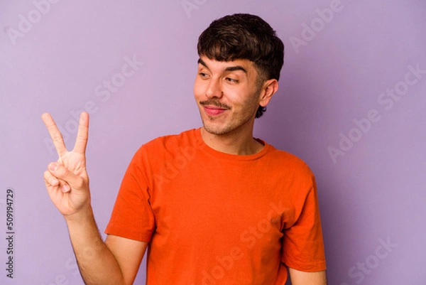 Fototapeta Young caucasian man isolated on purple background joyful and carefree showing a peace symbol with fingers.