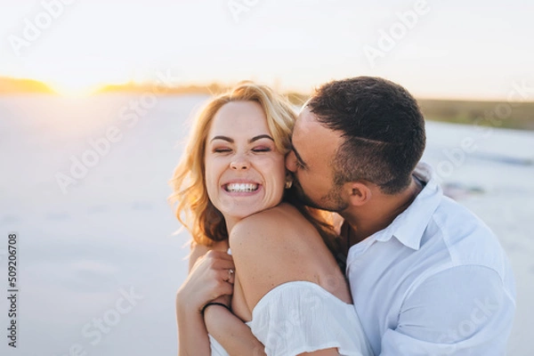 Obraz A tanned caucasian bearded guy in a white shirt kisses a young beautiful cheerful smiling blonde against the backdrop of the setting sun. Desert, sandy beach, rest and relaxation. Honeymoon concept.