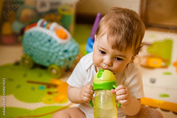 Fototapeta Cute little boy drinking water from a training bottle with a straw, home interior, lifestyle