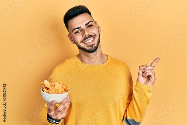 Fototapeta Young hispanic man with beard holding nachos potato chips smiling happy pointing with hand and finger to the side