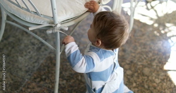 Fototapeta Cute baby toddler learning to stand up by leaning on chair furniture