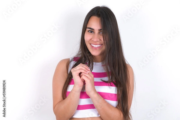 Fototapeta Dreamy charming young beautiful brunette woman wearing striped top over white wall with pleasant expression, keeps hands crossed near face, excited about something pleasant.