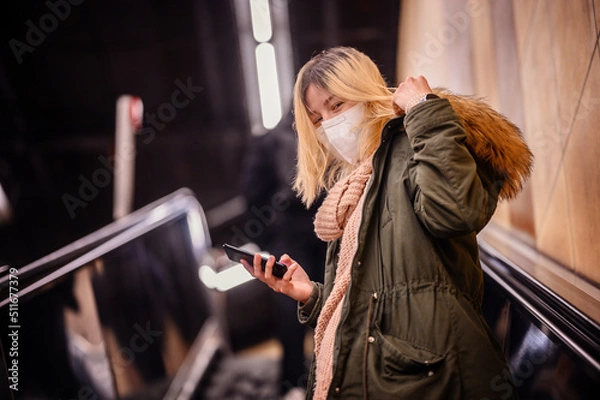 Fototapeta Blonde young woman in winter outfit and protective mask using smartphone going up the escalator