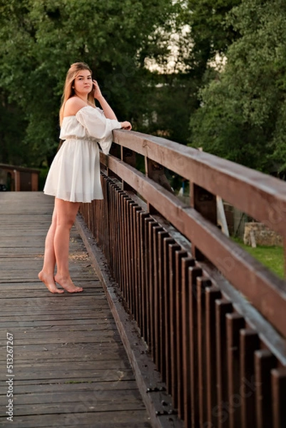 Fototapeta a beautiful girl in a white dress stands on a wooden bridge.