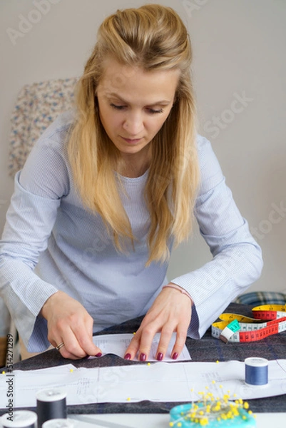 Fototapeta Young seamstress pinning up paper pattern on fabric. Woman tailor sews clothes with her own hands