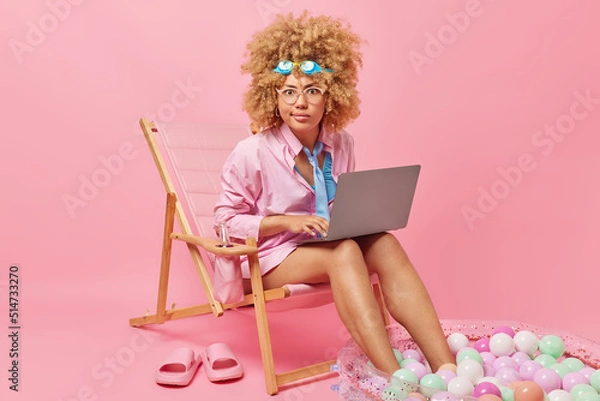 Fototapeta Serious curly haired businesswoman works online via portable computer poses on deck chair during summer vacation focused attentively at camera wears formal clothing isolated over pink background