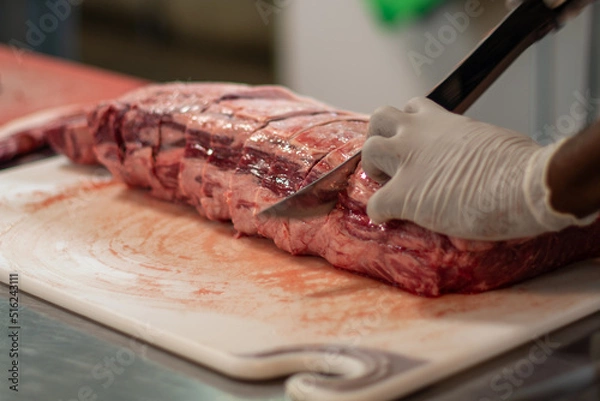 Fototapeta A close up of a large prime rib roast which is sitting on a brown plastic cutting board. The meat has some marbling and multiple ribs. The chef is using a long knife to cut steaks from the fresh meat.