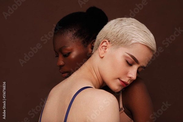 Obraz Two young women with different skin color standing together. Females put their heads on each other's shoulders with closed eyes.