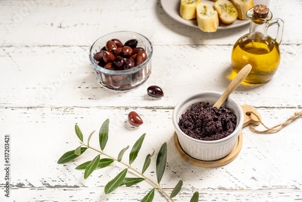 Fototapeta Olive pate in ramequin bowl and basic ingredients - olive oil, olives on white wooden background with crostini and olive branch