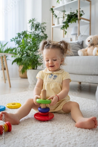 Obraz Vertical view of the female kid toddler playing wooden toys at home. Cute little kid sitting on the floor with toy eco-friendly pyramid in natural playroom and having fun