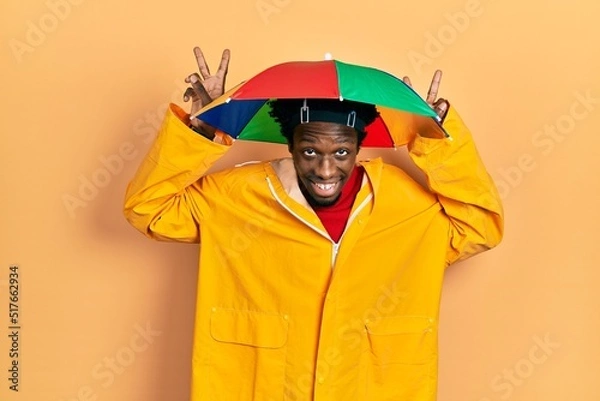 Fototapeta Young african american man wearing yellow raincoat posing funny and crazy with fingers on head as bunny ears, smiling cheerful