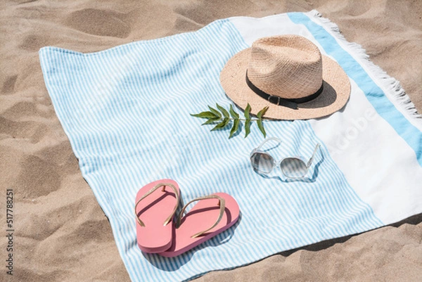 Fototapeta Beach towel with straw hat, sunglasses, leaves and flip flops on sand