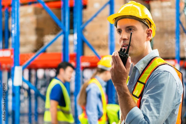 Obraz Warehouse worker working in factory warehouse industry and using radio talking communication, Foreman in hardhat safety vest with Two-Way radio working in logistics center