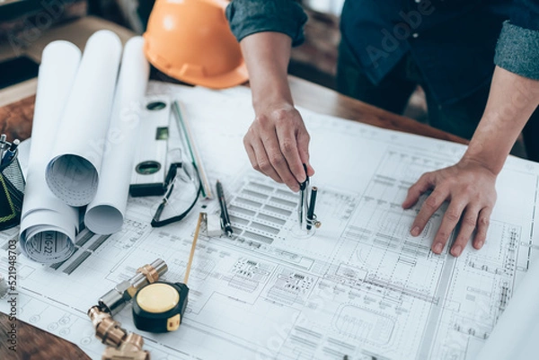 Fototapeta Engineering working with drawings inspection and writing on the office desk and Calculator, triangle ruler, safety glasses, compass on Blueprint. Engineer, Architect, Industry and factory concept.