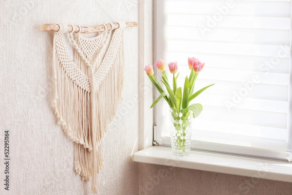 Fototapeta Handmade macrame. 100% cotton wall decoration with wooden stick hanging on a white wall.   Macrame braiding and cotton threads.  Female hobby.  ECO friendly modern knitting concept in the interior