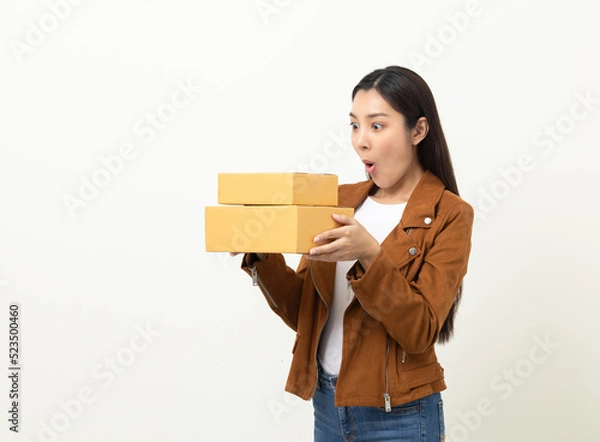 Fototapeta Young excited beautiful asian woman with many parcel cardboard standing on isolated white background. Cheerful female holding lot of parcel box receive from the delivery service