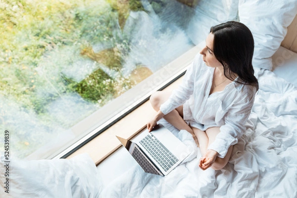 Fototapeta A young woman woke up and sitting with a laptop on a white bed, looking out the panoramic window. Calm morning. Remote work, social distance, freelance concept.