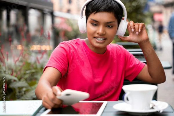 Fototapeta Beautiful smiling asian woman using mobile phone, shopping online sitting in cafe. Stylish Indian hipster female wearing wireless headphones listening music outdoors. Modern technology concept