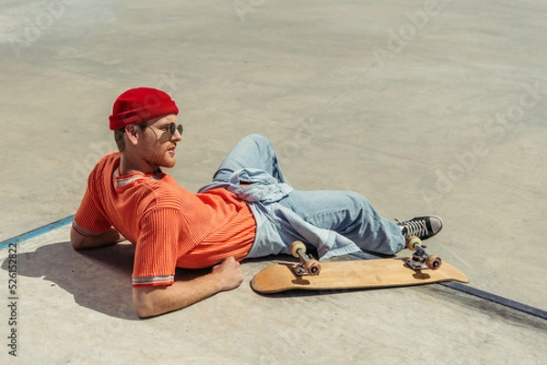 Fototapeta young man in orange t-shirt and beanie resting near skateboard in park