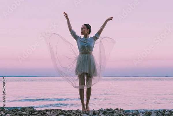 Fototapeta A young beautiful woman in a white dress fluttering in the wind, standing in the water.A happy free woman with her arms outstretched enjoys her rest.