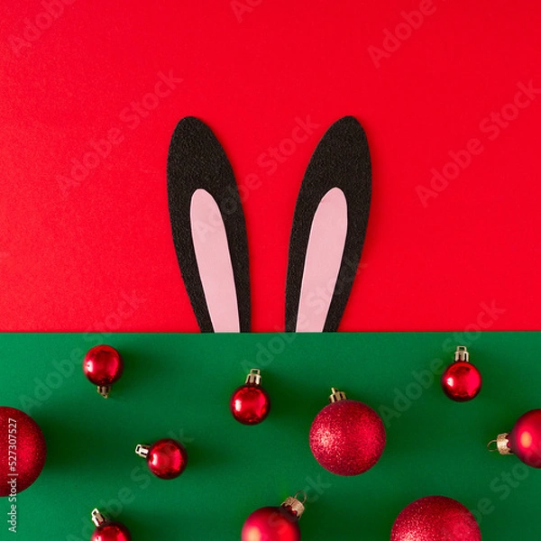 Fototapeta Creative flat lay holiday background made with Black rabbit ears like symbol of new year 2023 and red christmas balls on green background. Minimal New Year or Christmas concept.