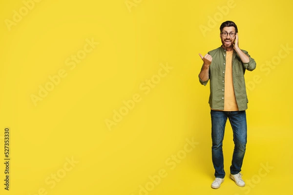 Fototapeta Wow offer. Excited shocked man pointing finger aside at free space for text posing over yellow studio background