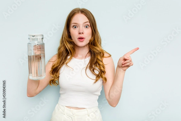 Fototapeta Young caucasian woman holding jar of water isolated on blue background pointing to the side